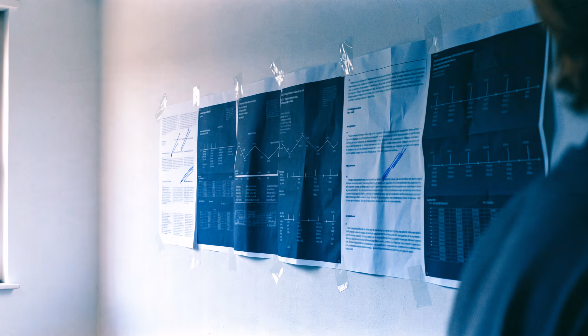 A printed project schedule spanning several taped sheets on an office wall, annotated by hand, with a team member working alongside it.